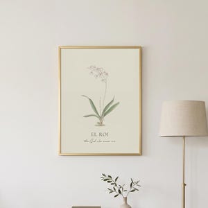 以下が含まれることがあります： 白い蘭の花と緑の葉の植物画が描かれた額装されたウォールアートプリント。花の下には「EL ROI」および「the God who sees me」というテキストが記されています。プリントは白い壁に細い金色のフレームで飾られています。
