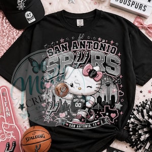 May include: Black t-shirt featuring a graphic of Hello Kitty in a Spurs basketball uniform, holding a basketball. The shirt has the words "San Antonio Spurs" and "San Antonio, TX". A black baseball cap with the Spurs logo is also visible.