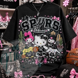 May include: Black t-shirt featuring the word "SPURS" in a stylized design with pink accents and cartoon characters. The shirt includes the text "SAN ANTONIO" and basketball-themed graphics. Accessories include a black baseball cap and a basketball.