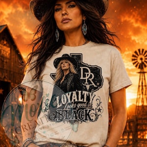 May include: A beige t-shirt with a graphic featuring a woman in a cowboy hat, the Texas outline, and the text "Loyalty Looks Good in Black." The shirt is worn by a person wearing a cowboy hat and jewelry.