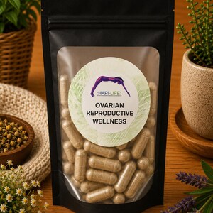 Puede incluir: Una bolsa negra con una ventana transparente que muestra numerosas cápsulas beige. La bolsa presenta una etiqueta circular con el texto "HAPI-LIFE: OVARIAN REPRODUCTIVE WELLNESS" y un gráfico morado. La imagen está sobre una superficie de madera con plantas decorativas.