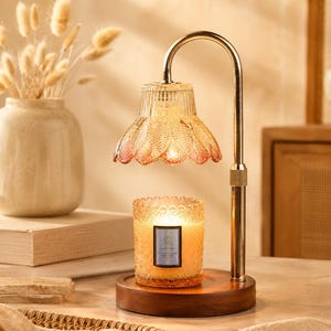 Op de afbeelding: Een kaarsverwarmerlamp met een goudkleurige metalen arm en een houten voet. De lamp heeft een geribbelde, amberkleurige glazen kap met een bladvormig patroon. Een cilindrische glazen kaars met een getextureerde rand staat op de voet.