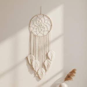 May include: A cream-colored macrame dream catcher with a circular design and hanging leaf accents. The wall decor is displayed against a neutral wall, with a woven basket and blanket in the foreground. A wooden shelf holds a vase and books.