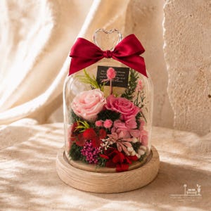 May include: A glass cloche with a heart-shaped handle, topped with a deep red satin bow, containing preserved pink and red roses, green moss, and small floral accents. A small black rectangular tag with white text is tucked inside the arrangement.