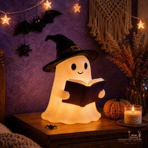 Könnte beinhalten: Eine leuchtende, weiße Lampe in Geisterform, die einen schwarzen Hexenhut mit violettem Band und goldener Schnalle trägt. Der Geist hat schwarze Augen und ein kleines Lächeln und hält ein offenes schwarzes Buch. Die Lampe strahlt warmes Licht aus.
