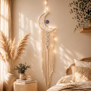 Puede incluir: Un tapiz de macramé en forma de media luna hecho de cuerda color hueso, con un patrón central de diamantes tejidos, flecos largos y un amuleto de ojo turco azul y blanco suspendido, iluminado por luces de cadena cálidas integradas.