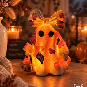 Puede incluir: Una figura de fantasma naranja brillante con un lazo grande en la cabeza, dos ojos negros circulares y un patrón de parches en el cuerpo. El fantasma sostiene un pequeño cubo de calabaza en una mano y la figura está iluminada desde el interior.