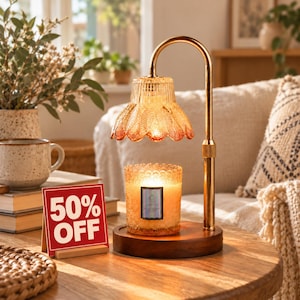 May include: A gold-toned metal candle warmer lamp with a scalloped amber glass shade and a wooden base, featuring a lit candle inside a textured glass jar. A red sign with white text reads 50% OFF, placed next to the lamp on a wooden table.
