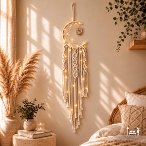 May include: A crescent moon-shaped macrame wall hanging made of off-white cord, featuring an integrated string of warm LED lights, a small wooden owl cutout, and intricate knotted patterns with long fringe tassels hanging vertically.