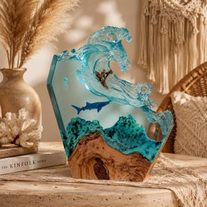 May include: A decorative resin and wood sculpture featuring a translucent blue ocean wave crashing over a natural wood base. A small surfer figure rides the wave, while a blue whale shark and a jellyfish are embedded within the clear resin.