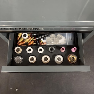 May include: A gray U.S. General tool drawer containing an assortment of welding consumables. The drawer features a black foam organizer holding various ceramic nozzles, brass contact tips, and metal components, with extra parts in a plastic bag.