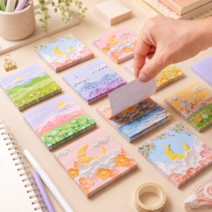 Op de afbeelding: Een verzameling vierkante memoblaadjes met getextureerde landschapsillustraties in impasto-stijl, waaronder maansikkels, bloemenvelden, bergketens en wolken, waarbij één blaadje wordt opgetild om een lege witte onderkant te onthullen.