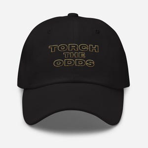 May include: A black baseball cap featuring the text TORCH THE ODDS embroidered in gold thread across the front. The lettering has a distinct horizontal line texture within each character, set against the solid black fabric of the cap.
