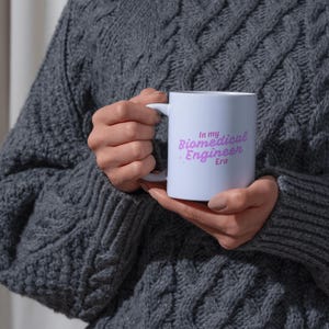 May include: A white ceramic mug with the text "In my Biomedical Engineer Era" in a purple script. The mug has a standard handle and is held by a person wearing a gray cable-knit sweater. The mug is a perfect gift for biomedical engineers.