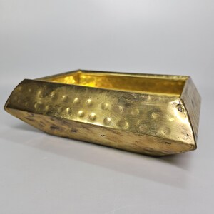 May include: A rectangular gold-colored metal tray with a hammered texture on the exterior sides. The vessel features angled edges and a smooth, reflective interior surface, creating a shallow basin with a metallic finish and visible indentations.