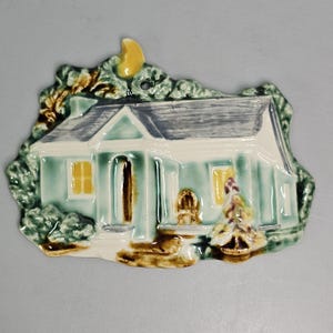 May include: A ceramic wall plaque shaped like a house with a light blue exterior, a grey roof, and yellow windows. The design includes a yellow crescent moon at the top, green foliage surrounding the structure, and a small brown entryway.