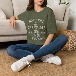 May include: An olive green short-sleeve t-shirt featuring a graphic of a small dragon holding a book. The text reads "DON'T KILL MY CREATURES I'LL CRY" with five circular icons representing different elements arranged above and below the dragon.