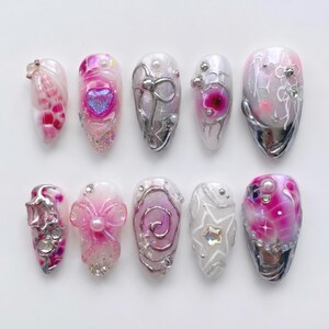 Könnte beinhalten: Ein Set aus zehn künstlichen Fingernägeln mit verschiedenen Designs in Rosa-, Weiß- und Silbertönen. Die Nägel sind mit 3D-Elementen verziert, darunter metallische silberne Wirbel, schillernde Strasssteine, Perlen, Schleifen und Sternformen.