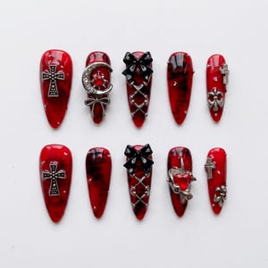 Peut inclure: Un ensemble de dix faux ongles en forme d'amande avec une base marbrée rouge et noire brillante. Les ongles sont ornés de breloques en métal argenté, notamment des croix, un croissant de lune, des nœuds, un cœur rouge et des strass.