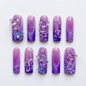 Könnte beinhalten: Ein Set aus zehn künstlichen Fingernägeln mit violettem Farbverlauf und handgemalten weißen Wirbel- und Schmetterlingsmustern. Mehrere Nägel sind mit dreidimensionalen, transparenten Bärenanhängern, kleinen violetten Perlen und Sternen verziert.
