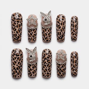Könnte beinhalten: Ein Set aus zehn künstlichen Nägeln mit Leopardenmuster. Einige Nägel weisen silberfarbene Hasen- und Sternanhänger mit Strasssteinen auf. Die Nägel sind quadratisch und vor einem weißen Hintergrund angeordnet.