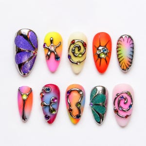 Könnte beinhalten: Ein Set aus zehn künstlichen Fingernägeln mit verschiedenen Designs, darunter Farbverläufe, Glitzertexturen und metallisch silberne Auflagen. Jeder Nagel ist mit kleinen, bunten, eingebetteten Strasssteinen in Wirbel- und Blumenmustern verziert.
