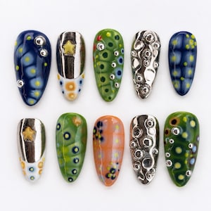 Könnte beinhalten: Ein Set aus zehn künstlichen Fingernägeln mit verschiedenen Designs, darunter dunkelblau mit gelben Punkten, metallic-silber mit gelben Sternen, grün mit bunten Punkten und silbernen Nieten sowie texturierte silberne Oberflächen mit Kreismustern.
