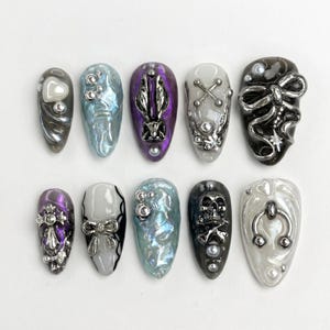 Pode incluir: Um conjunto de dez unhas postiças com vários designs, incluindo berloques metálicos prateados, laços, cruzes, caveiras e texturas iridescentes. As unhas estão dispostas em duas fileiras, exibindo uma mistura de paletas de cores preta, branca, roxa e azul.