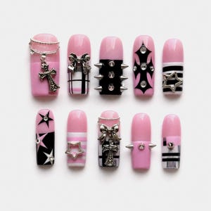 Könnte beinhalten: Ein Set aus zehn künstlichen Fingernägeln in Rosa und Schwarz. Die Designs umfassen silberne Kreuzanhänger, metallische Schleifen, spitze Nieten, schwarz-weißes Karomuster, horizontale Streifen sowie sternförmige Metallakzente mit eingelassenen Kristallen.
