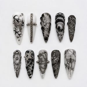 May include: A set of ten almond-shaped press-on nails featuring a monochromatic black, white, and silver color palette. The designs include metallic butterfly charms, studded crosses, silver chains, star-shaped rhinestones, and marbled patterns.