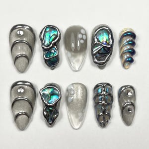 Könnte beinhalten: Eine Sammlung von zehn künstlichen Nägeln mit verschiedenen Designs. Einige Nägel sind silberfarben mit einer abgerundeten Form, andere weisen schillernde Abalone-Muscheln auf, und einige haben einen Farbverlauf. Die Nägel sind auf einer weißen Oberfläche angeordnet.