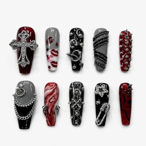 May include: A set of ten press-on nails featuring a coffin shape with various designs including a silver cross, red lip prints, silver chains, metal studs, a star charm, a safety pin, and marbled red and black patterns in a dark color palette.