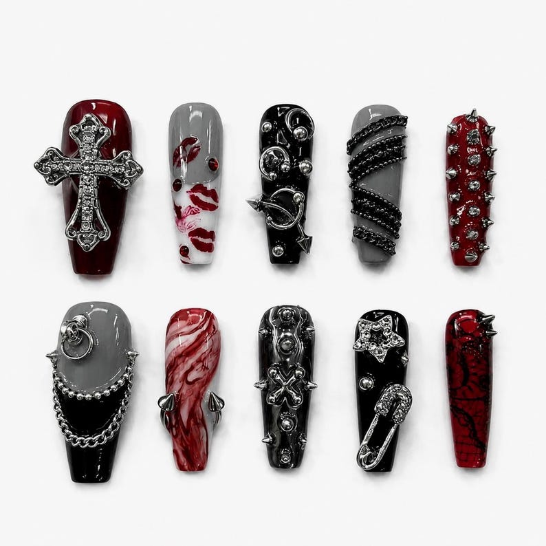 May include: A set of ten press-on nails featuring a coffin shape with various designs including a silver cross, red lip prints, silver chains, metal studs, a star charm, a safety pin, and marbled red and black patterns in a dark color palette.