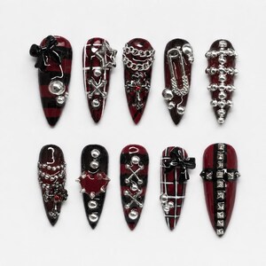 Könnte beinhalten: Ein Set aus zehn mandelförmigen Press-on-Nägeln in Dunkelrot und Schwarz. Die Designs umfassen Karomuster, schwarze Schleifen, silberne Ketten, Sicherheitsnadeln, Metallnieten, Kreuzanhänger und herzförmige Akzente in einem stimmigen Set.