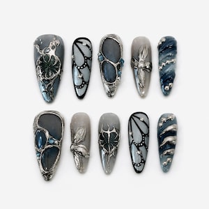 Könnte beinhalten: Ein Set von zehn künstlichen Nägeln in verschiedenen Designs. Jeder Nagel hat eine andere Form und weist eine Kombination aus silbernen Metallakzenten, Blau- und Grautönen sowie dekorativen Elementen wie Blumen und Schmetterlingsmustern auf.