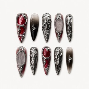 Könnte beinhalten: Ein Satz von zehn langen, spitz zulaufenden Kunstnägeln. Jeder Nagel hat ein silbernes Metallic-Design mit roten, schwarzen und grauen Akzenten. Die Designs variieren, einschließlich verzierter Muster und Farbverläufe.