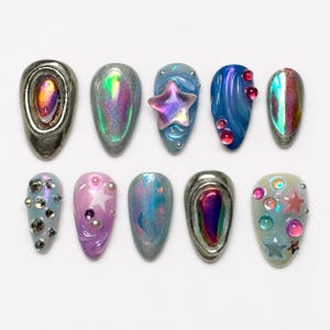 Könnte beinhalten: Ein Set aus zehn künstlichen Fingernägeln mit verschiedenen Designs, darunter irisierende Oberflächen, metallisch silberne Ränder, erhabene Sternformen sowie eingebettete bunte Strasssteine und Perlen in Blau, Lila, Silber und Rosa.