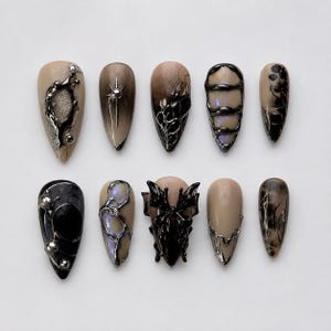 Könnte beinhalten: Eine Sammlung von zehn mandelförmigen Kunstnägeln. Die Nägel weisen eine Vielzahl von Designs auf, darunter metallische Akzente, komplizierte schwarze Muster und ein zentrales schwarzes, skulpturales Element. Farben sind Beige, Schwarz, Silber und schillernde Farbtöne.