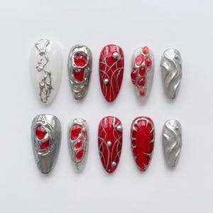 Könnte beinhalten: Ein Set aus zehn künstlichen Fingernägeln in Mandelform mit einer Kombination aus roten, weißen und silbernen Metallic-Designs. Die Nägel zeigen erhabene silberne Flüssigmetall-Texturen, rote durchscheinende Edelsteine und feine Linien.