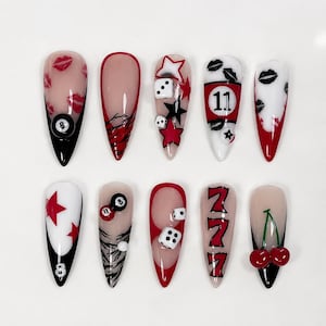 May include: A set of ten long, almond-shaped artificial nails. Each nail features unique designs, including red lips, dice, stars, the number 11, and cherries. The nails have a glossy finish with black, red, and white accents.