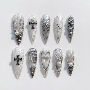 Könnte beinhalten: Ein Set aus zehn langen, spitz zulaufenden Kunstnägeln. Jeder Nagel hat eine weiße Basis mit aufwendigen silbernen Designs, darunter Kreuze, Schmetterlinge und abstrakte Muster. Einige Nägel sind mit Perlen und metallischen Akzenten verziert.