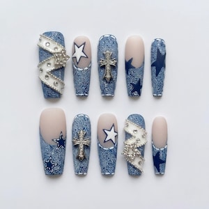 Könnte beinhalten: Ein Set aus zehn künstlichen Fingernägeln mit blauem Jeans-Druck, weißen Sternen, silbernen Kreuz-Anhängern und weißen, gewellten Bändern, die mit kleinen klaren Strasssteinen besetzt sind. Die Nägel haben matte Nude- und Jeans-Texturen.