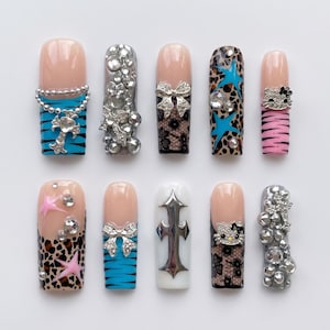 Peut inclure: Un ensemble de dix faux ongles présentant des motifs variés, notamment des rayures tigrées bleues avec une croix argentée, des strass, de la dentelle noire, un imprimé léopard avec des étoiles, des rayures zébrées roses, des motifs Hello Kitty.
