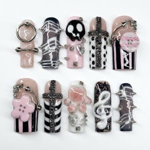May include: A set of ten press-on nails featuring diverse designs including a silver ring, black and white stripes, a skull, a cross, a pink flower, a treble clef, and metal spikes. The nails are decorated with various textures, charms, and patterns.