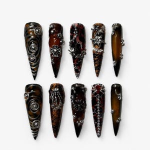 Könnte beinhalten: Ein Set aus zehn stilettoförmigen Press-on-Nägeln mit einer dunkelbraunen und schwarzen Schildpatt-Basis. Jeder Nagel ist mit silberfarbenen Metallanhängern verziert, darunter Kreuze, Ketten, Kugeln, Spinnenmotive und skelettartige Handakzente.