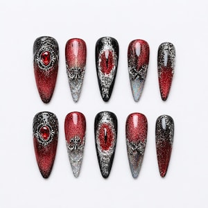 Könnte beinhalten: Ein Set aus zehn stilettoförmigen künstlichen Nägeln mit einem dunkelroten und schwarzen Glitzerverlauf. Die Designs umfassen silberne filigrane Verzierungen, ein zentrales rotes Drachenaugenmotiv, kleine rote Strasssteine und gekreuzte silberne Schwerter.