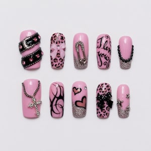 Könnte beinhalten: Ein Set aus zehn künstlichen Fingernägeln mit hellrosa Grundfarbe. Die Designs umfassen einen schwarzen Gürtel mit rosa Nieten, Leopardenmuster mit rosa Schleife, eine silberne Sicherheitsnadel, den Schriftzug I love you, schwarze Perlen, ein Kreuz-Anhänger, Tribal-Muster, Herzen und Schleifen.