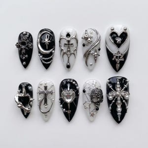 Könnte beinhalten: Ein Set aus zehn künstlichen Fingernägeln in Schwarz und Weiß mit metallisch silbernen Akzenten. Die Designs enthalten komplexe 3D-Elemente wie Kreuze, Herzen, Sterne, Ketten, Perlen und Totenkopfmotive auf mandelförmigen Nagelspitzen.