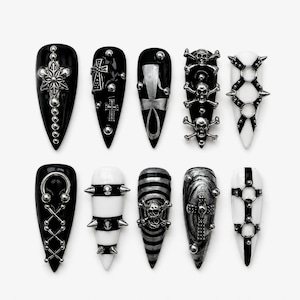Könnte beinhalten: Ein Set aus zehn stilettoförmigen künstlichen Fingernägeln mit schwarzer und weißer Grundfarbe. Die Designs enthalten silberfarbene Metallverzierungen wie Totenköpfe, Kreuze, Ankh-Symbole, Nieten, Stacheln, Ringe und überkreuzte Muster.