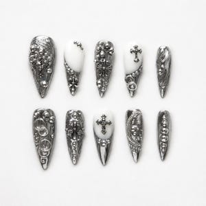 Könnte beinhalten: Ein Set aus zehn stilettoförmigen Press-on-Nägeln mit metallisch silbernem Finish, weißen Akzenten und dreidimensionalen Verzierungen wie silbernen Kreuzen, Kugeln, Ringen und komplexen, strukturierten Mustern auf jedem Nagel.
